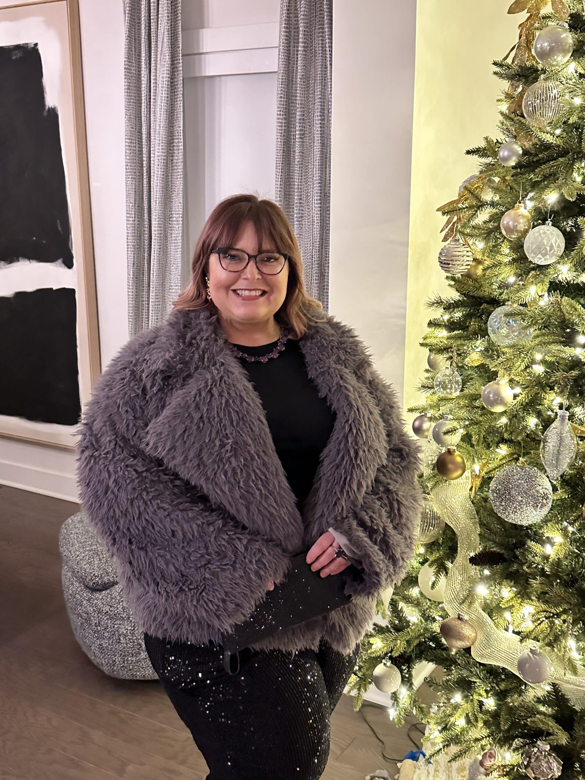 Kelly, co-founder of The Kel-Squared Collective, smiling next to a decorated Christmas tree. She is wearing a grey faux fur jacket and black sequined pants. Organizer of The Grown-Ups Table events in Sylvania, Ohio.